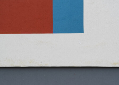 Abstract graphic view of a stucco building wall highlighting the intersection of bright red, blue, white, and dark grey painted sections.