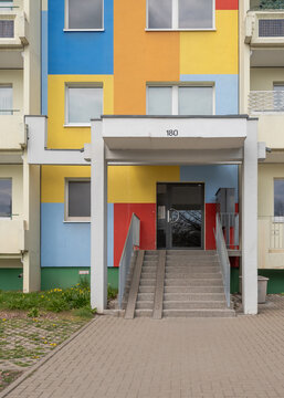 Colorful modern entrance to a residential apartment block numbered 180 featuring a concrete canopy, stairs with a ramp, and a vibrant blue, yellow, and red patchwork facade.