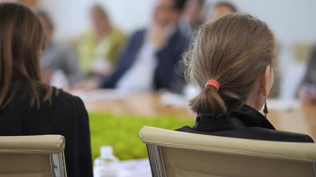 Woman with ponytail sits at conference table. Russian flag beside her. Blurred attendees in background. Professional meeting atmosphere. Suitable for business, diplomacy, or international negotiations