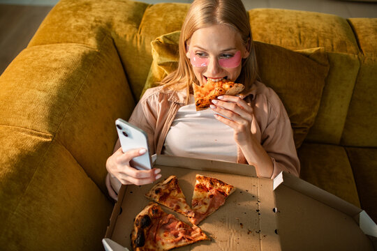 Young woman eating pizza on sofa at home with smartphone and eye patches