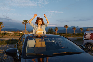 Fototapeta premium Young woman enjoys a joyful moment through an open car sunroof in a desert setting, casual denim jacket, striped top, warm sunset light, free spirited lifestyle
