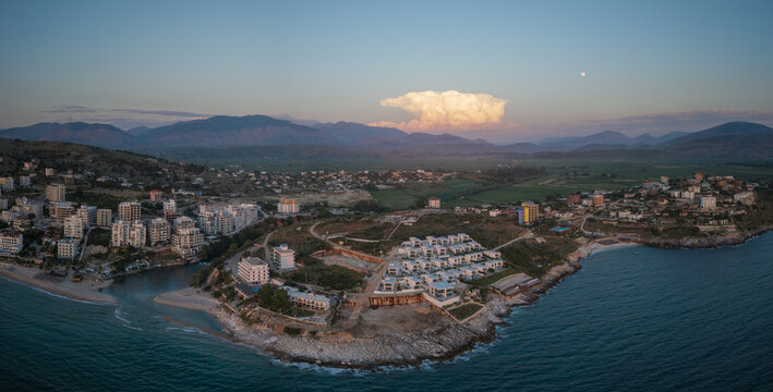 Aerial view of Saranda Beach and the coastal town of Sarande with modern buildings, rocky shoreline, and distant mountains at sunset in Sarande, Vlore County, Albania.