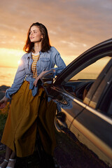 Fototapeta premium Cheerful woman outdoors beside a car at sunset, denim jacket over striped shirt, yellow skirt, relaxed pose, warm light, natural colors, airy mood