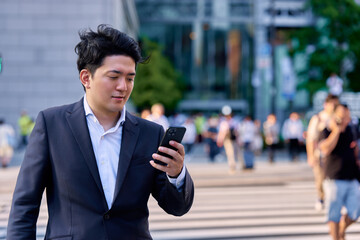 人々の行き交う夕方の都会のオフィス街の交差点の雑踏の中でスマートフォンを操作するビジネスマンの男性 © Hiroba Studio