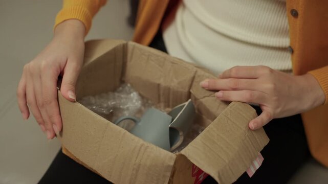 Close-up Stressed young woman opening a parcel and discovering damaged product after online shopping at home. Shipping damage, delivery issue. E-commerce, consumer rights and insurance claim concept.
