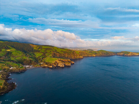 Aerial view of Farol do Topo lighthouse on the rugged coastline with green hills, dramatic cliffs, and the deep blue Atlantic Ocean in Topo, Acores, Portugal.