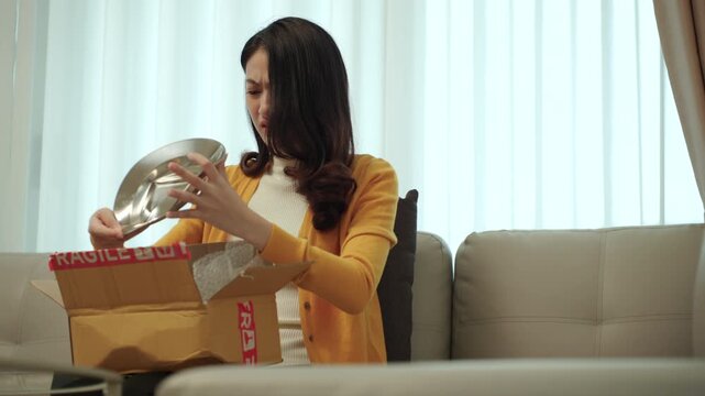 Stressed young woman opening a parcel and discovering damaged product after online shopping at home. Shipping damage, delivery issue. E-commerce, consumer rights and insurance claim concept.
