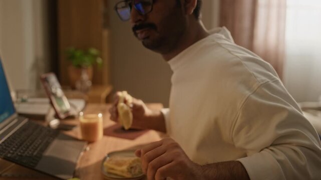 Zoom out shot of Indian young man typing on laptop while eating sandwich at home office desk. Footage represents remote work, freelance lifestyle, and flexible working hours concepts