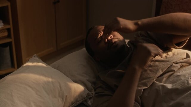 High angle shot of young African American man waking up in bed, checking smartphone while squinting and rubbing eyes. Footage ideal for sleep disorder awareness or digital detox campaigns