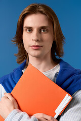 Obraz premium Stylish young student with long hair holding bright orange folder against blue backdrop