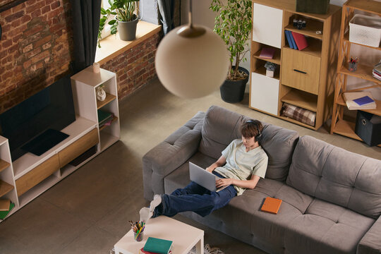 Teen boy studying with laptop and headphones on sofa in modern living room. Concept of online learning, remote school, home education, digital skills and productivity.