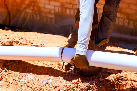 Worker holds white pipe and uses tool to cut it while on dirt surface at construction site.