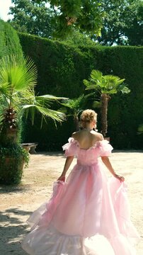 Beautiful Caucasian woman in a luxurious pink tulle dress spinning and walking through a lush green park with palm trees on a bright sunny day, slow motion.