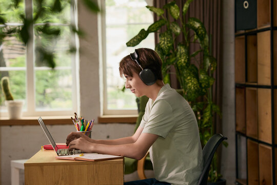 Teen boy typing on laptop with headphones in green home workspace. Concept of online education, remote school, focused study, digital skills and productivity.