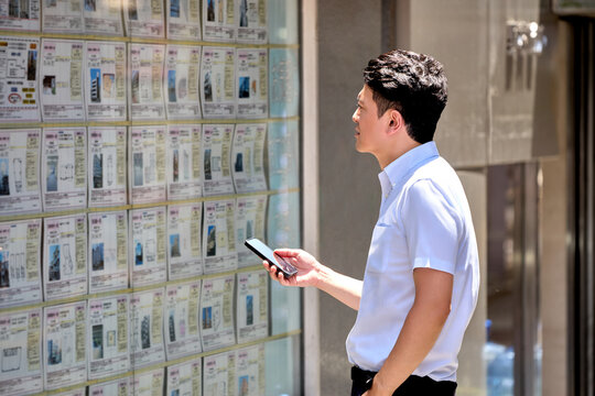 スマートフォンを持って不動産の賃貸や物件情報の広告を店頭の掲示板で見ている30代の日本人の男性