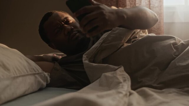 Medium shot of young African American man waking up in bed, checking smartphone, and covering head with pillow. Ideal for sleep health, stress management, or digital detox campaigns