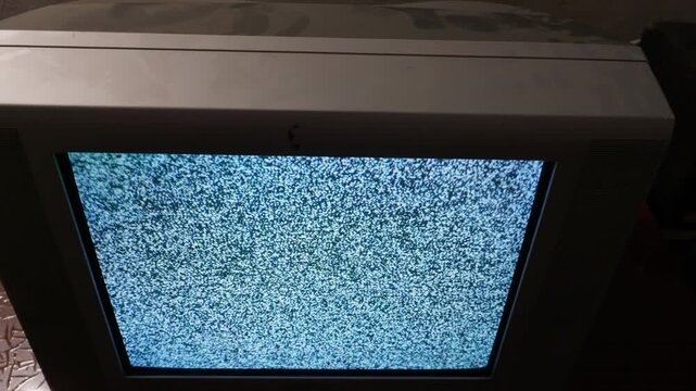 An old cathode ray tube television rests on a dark wooden surface inside a dimly lit room. The curved glass screen displays black and white static noise. Dust covers the top casing.