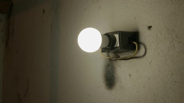 A single round light bulb is mounted on a dark, rough-textured concrete wall in a dimly lit industrial space. Concept of isolation, darkness, industrial decay, and a single spark of hope.