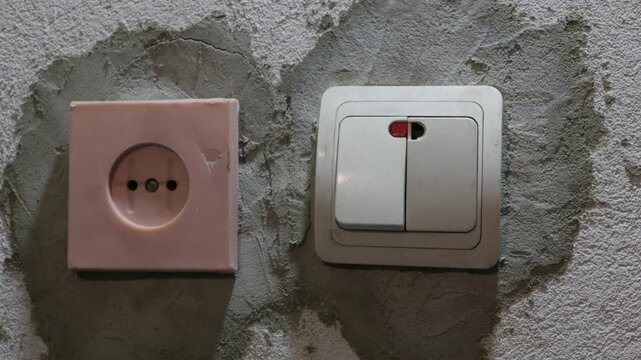 A male hand presses down on a double white light switch located next to an old power socket on a coarse, unfinished plaster wall in dim lighting. Concept of energy saving and renovation.