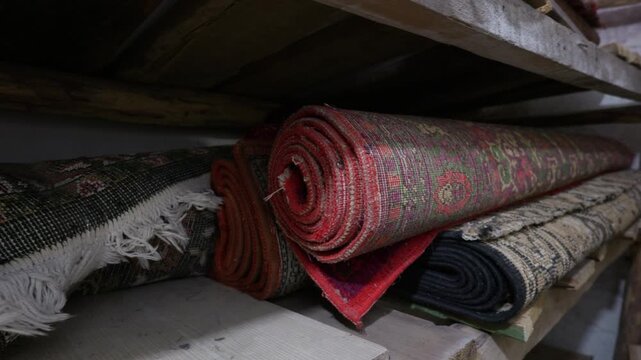 Traditional patterned carpets rest rolled up and stacked on rustic wooden shelves. The ornate woven rugs feature colorful designs and classic fringe edges. They sit in a dim storage room.