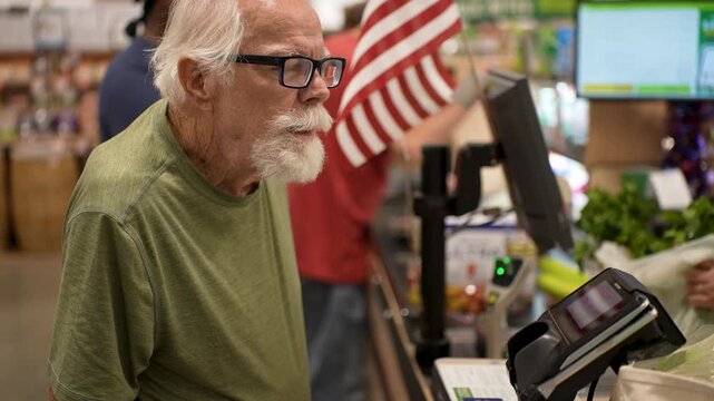 Senior man in a grocery store smiles as he checks out his items and uses a credit card at the counter with a cashier