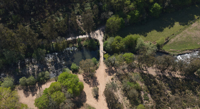 Aerial view of a person crossing a stone bridge over a river in a lush park with surrounding trees and dirt paths in Valongo, Porto District, Portugal.
