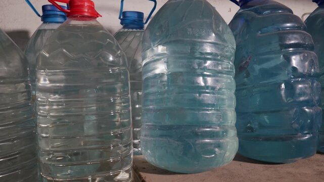 Large plastic water jugs and bottles sit on a dusty wooden shelf in a dark room. The containers vary in shape and color, with some filled with clear water and others with blue liquid.