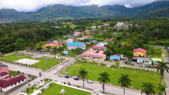 Aerial view of the rural town center of Ainaro Regency in Timor-Leste, featuring green parks, local buildings, and a stunning mountainous backdrop.