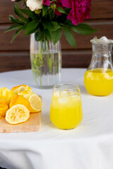 Fototapeta premium Bright still-life scene showing glass of cold yellow lemon beverage with ice, matching pitcher filled with juice, wooden cutting board with halved and peeled
