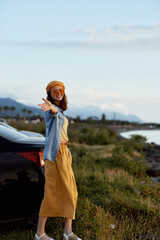 Fototapeta premium Woman lifestyle outdoors by coast, standing near car, sunny day, relaxed pose, pointing to camera, hat, denim jacket, mustard skirt, striped top, natural scenery, tranquil mood, travel vibes