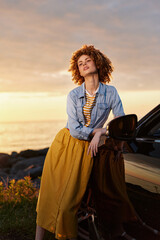 Fototapeta premium Relaxed woman with curly hair leaning against a car at sunset, wearing a denim shirt and mustard skirt. A candid lifestyle moment focused on fashion and outdoor confidence.