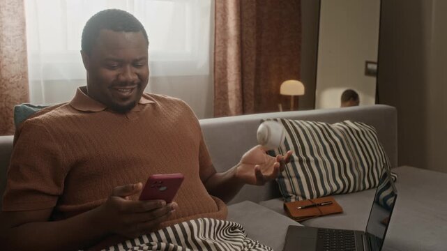 Medium shot of African American young man sitting on sofa, using smartphone and tossing baseball during remote work break. Clip suitable for promoting employee wellbeing and hybrid lifestyle