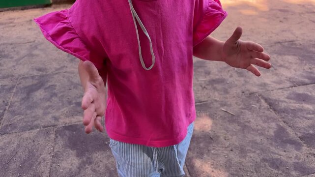 A small child in a pink T-shirt claps while playing outdoors.
