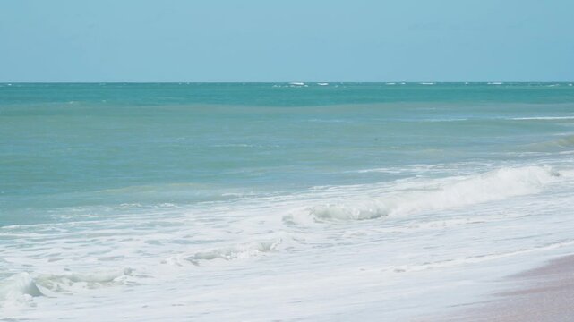 Blue sea meets light pink sand. Gentle waves roll toward shore. Clear sky above. White foam spreads across wet beach. Soft waves break near shore. Ideal for vacation, travel, and relaxation themes