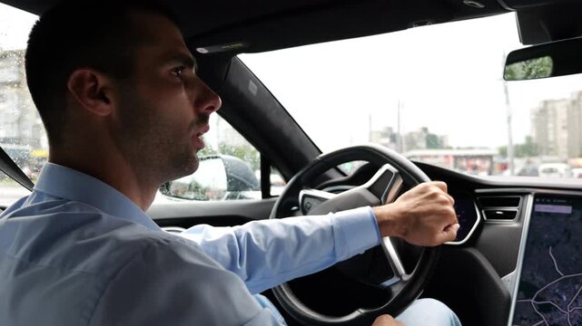 Handsome businessman driving a modern electric car at urban road. Male successful entrepreneur riding on electrical vehicle at rain city. Young man commuting to work at rainy day. Slow mo