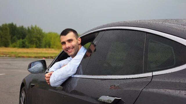 Handsome successful entrepreneur in shirt leaning out of driver window of modern electric car and looking into camera. Portrait of smiling businessman in innovative electrical vehicle at rainy day