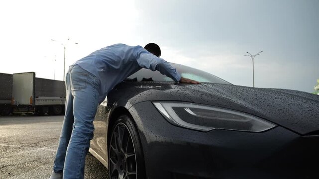 Successful smiling entrepreneur rejoices to purchase new electrical automobile at sunny rainy day. Happy young businessman in shirt enjoying modern electric auto. Concept of buy or rent car. Slow mo