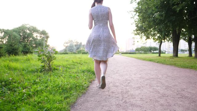 Young adult woman in summer dress stroll along park alley, camera follow behind. Slow motion shot, girl leisurely come forward, empty gravel path, green grass and trees around. Soft natural light