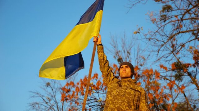 Male ukrainian army soldier lifting blue-yellow banner in honor of victory against russian aggression. Young military in camouflage uniform raised flag of Ukraine at countryside. Invasion resistance