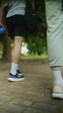 Two people walk on a paved path through a green park, seen from a low angle. Their legs and athletic shoes are in motion on the outdoor trail.