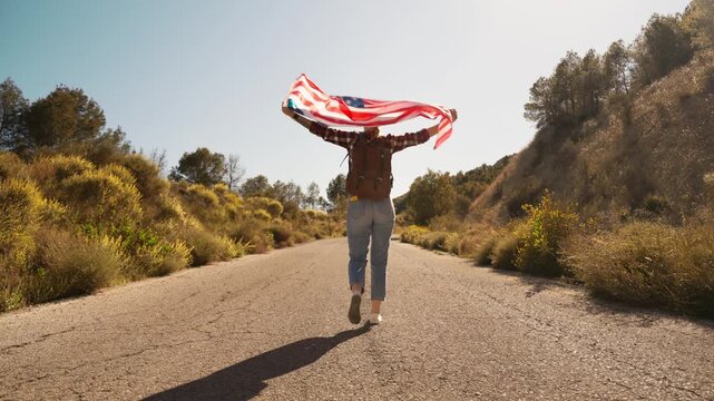 Young woman with backpack and USA flag hitchhiking on open road in scenic landscape, symbolizing freedom, independence and travel lifestyle.Open road, freedom lifestyle, independence concept