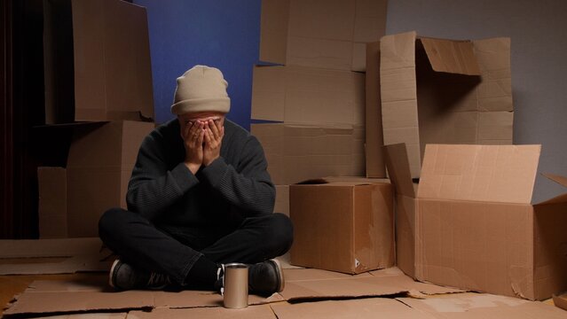 Homeless man experiencing despair and poverty, sitting on cardboard surrounded by boxes, hiding his face in his hands while begging for help with a metal can