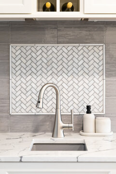 A prep or bar sink detail shot in a luxury kitchen with white herringbone and grey subway backsplash tiles, white marble countertop, and white cabinets.