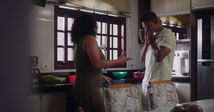 Husband responding with visible frustration as wife argues in kitchen, highlighting relationship tension and emotional dynamics during intense conflict, African descent