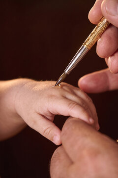 Orthodox Christian priest anointing baby hand with holy oil using golden ornate brush during baptism chrismation sacrament ceremony, close-up of religious ritual on dark background.

