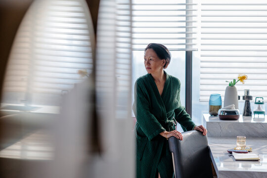 Woman in robe looks out window, thoughtful.