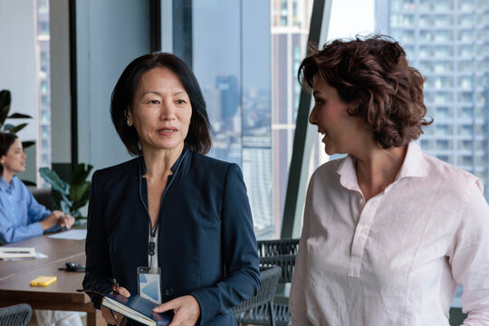 Businesswomen converse in a modern office with a city view.