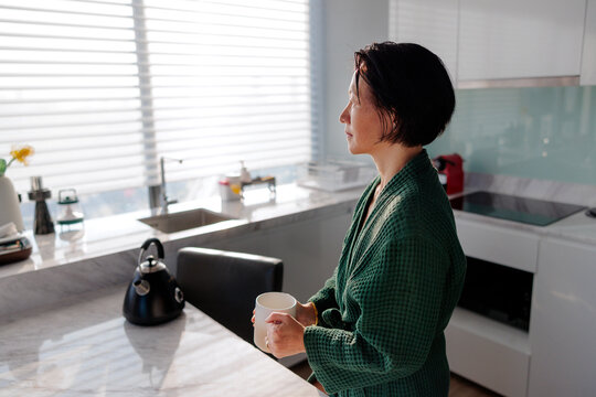 Woman in robe with mug looks out kitchen window.