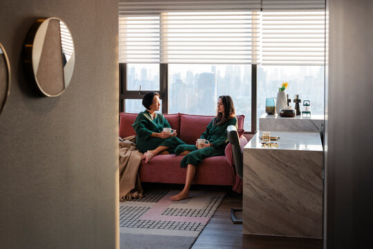 Two women in robes share coffee chat with city apartment view