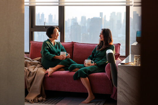 Two women share morning coffee in robes with city view from couch